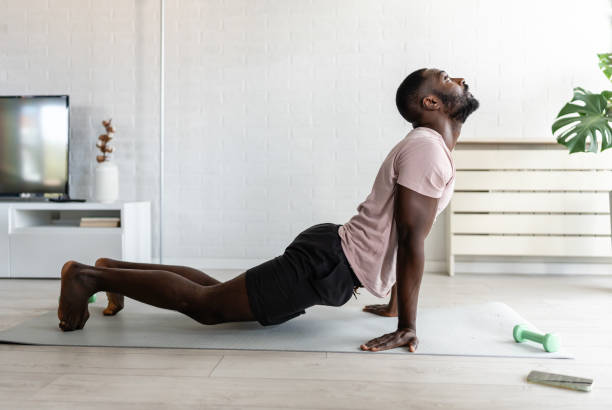 istock Young African-American man doing yoga on a mat in a bright home setting, practicing the cobra pose with focused posture. Surrounded by plants, promoting wellness and relaxation. 2181961592