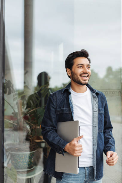 istock Smiling young man holding laptop walking near office building window 2181963762