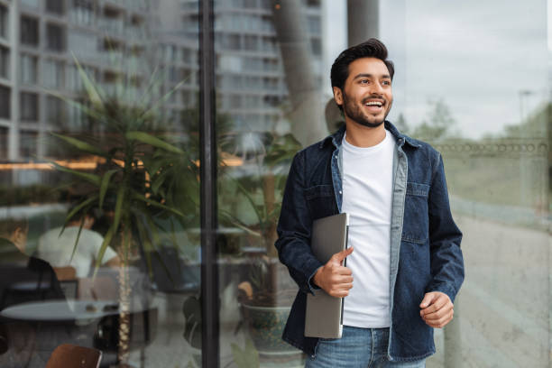 Smiling young man holding laptop walking near office building