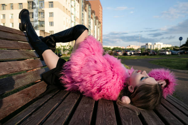 Woman in a pink fur coat and boots relaxing on a wooden deckchair in an urban green space. Stylish and modern, enjoying the sunlight.