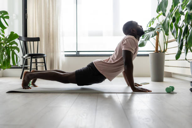 Young African-American man doing yoga on a mat in a bright home setting, practicing the cobra pose with focused posture. Surrounded by plants, promoting wellness and relaxation.