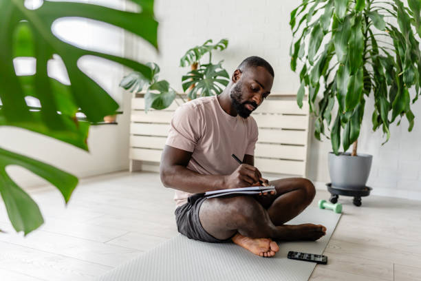 young african-american man sitting cross-legged on a yoga mat at home, writing in a notebook after a workout, with a smartphone nearby, creating a calm and focused atmosphere. - dagbok bildbanksfoton och bilder