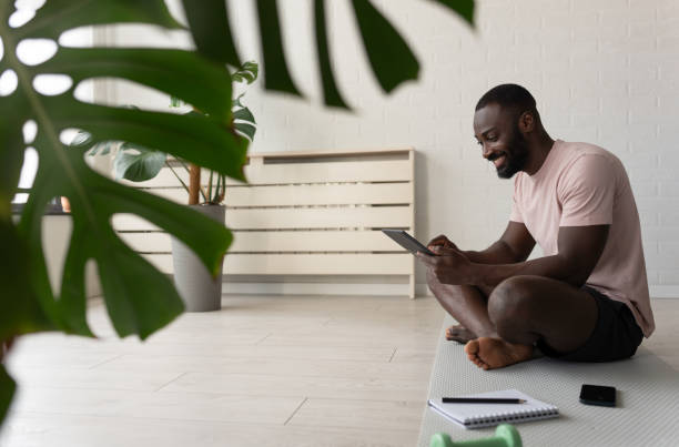 istock Young African-American man sitting on a yoga mat at home after exercising, smiling while using a tablet for a wellness app in a bright, plant-filled space. 2181970001