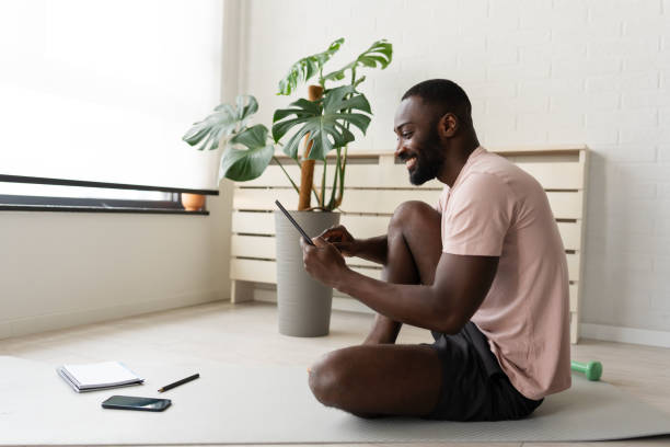 istock Young African-American man sitting on a yoga mat at home after exercising, smiling while using a tablet for a wellness app in a bright, plant-filled space. 2181970065