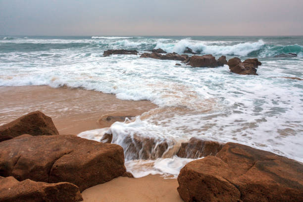 Waves gently cascade over large rocks on a sandy beach, creating a dynamic blend of motion and tranquility along the shoreline