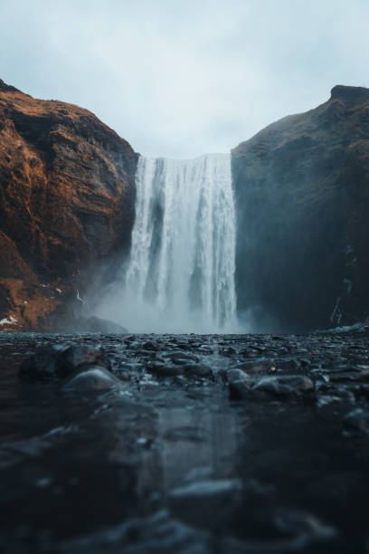A breathtaking view of Skogafoss, one of Iceland's most famous waterfalls, cascading down steep, rocky cliffs into a shallow pool. The scene is framed by dark, rugged terrain with a dramatic sky above, enhancing the atmosphere of solitude and natural beauty. The low-angle shot emphasizes the scale and raw power of Skogafoss, creating a sense of awe and tranquility in the Icelandic wilderness.