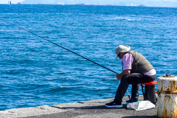 Castro Urdiales, Cantabria-Spain, September 19, 2024;\nA retired man is fishing on the seashore, he is sitting down and putting bait on his fishing rod.