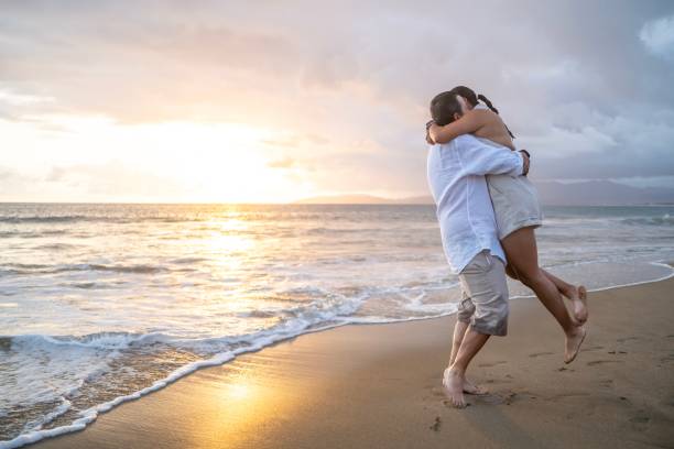 Happy couple on Hawaii vacation walking on beach with Hawaiian leis and Aloha clothing. Caucasian man wearing typical Hawaiian shirt and Asian woman girlfriend in pink sarong fabric sundress.