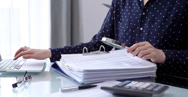 istock Businesswoman using calculator and reviewing ring folder of financial documents with magnifying glass at desk in modern office. Audit and taxes in business 2181996332