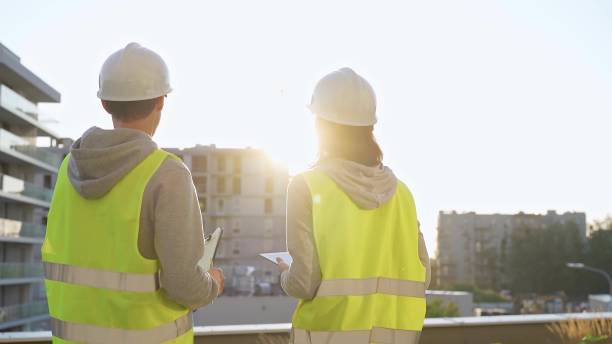istock Two engineers are standing on a construction site wearing hard hats and safety vests looking at a building and discussing something 2181997404