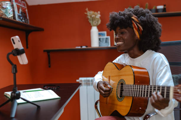 An enthusiastic woman, seated in a cozy room plays guitar while participating in an online music lesson, radiating joy and passion for learning and music.