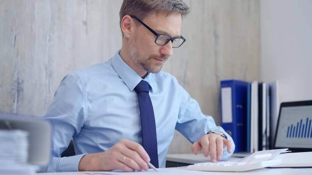 istock Male accountant with blue t-shirt concentrating while calculating costs and taking notes at his desk. Taxes, audit in business 2182002613