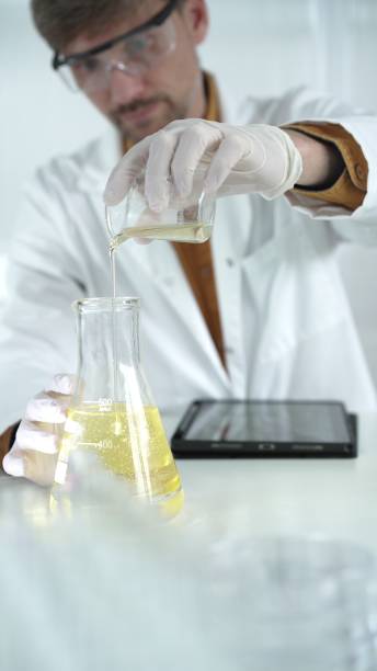 istock Man scientist with lab coat, white gloves, and protective glasses, is pouring a yellow oily liquid from one beaker to another near microscope in a laboratory, vertical close-up view 2182017567