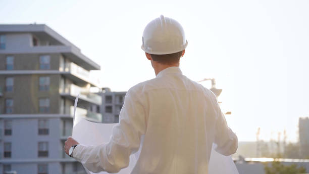 istock Construction site manager, wearing safety helmets and glasses, is holding blueprints and inspecting a building site during sunrise or sunset, back view. Architecture and engineering 2182018858
