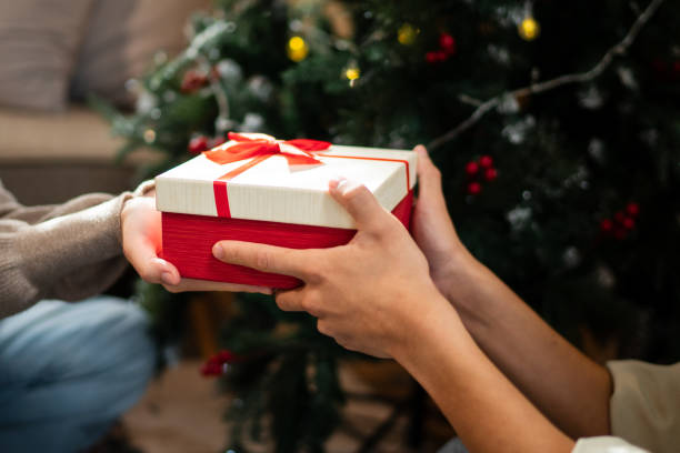 istock Two hands exchange a beautifully wrapped red and white gift box with a red bow, set against a backdrop of a decorated Christmas tree, creating a warm holiday moment 2182022832