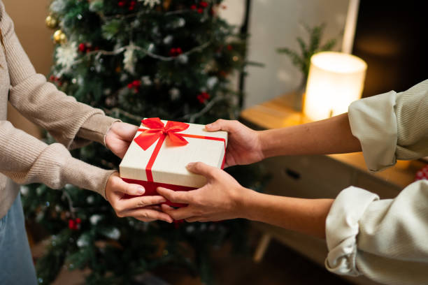 istock Two hands exchange a beautifully wrapped red and white gift box with a red bow, set against a backdrop of a decorated Christmas tree, creating a warm holiday moment 2182022840