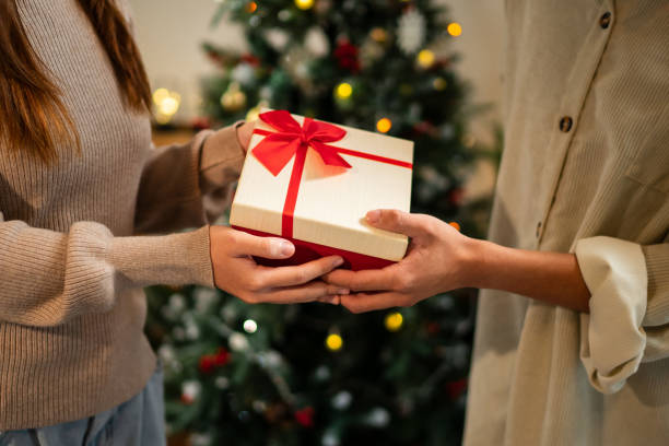 istock Two hands exchange a beautifully wrapped red and white gift box with a red bow, set against a backdrop of a decorated Christmas tree, creating a warm holiday moment 2182022856