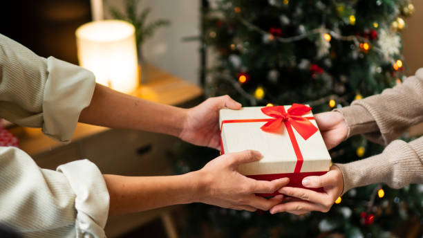 istock Two hands exchange a beautifully wrapped red and white gift box with a red bow, set against a backdrop of a decorated Christmas tree, creating a warm holiday moment 2182022857
