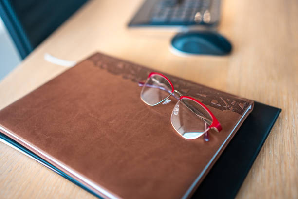 A pair of stylish glasses lies atop a closed brown leather notebook, with a computer mouse nearby, indicating a pause in a professional's workday within an office setting.