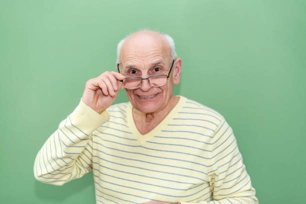 Old man smiling warmly, lifting his glasses while looking at camera against green background, wearing striped yellow sweater.