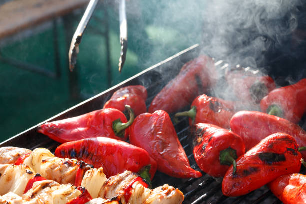 A close-up of a barbecue grill with skewers of grilled meat and vegetables alongside roasted red peppers. Smoke rises from the grill, enhancing the appeal of the freshly charred food.