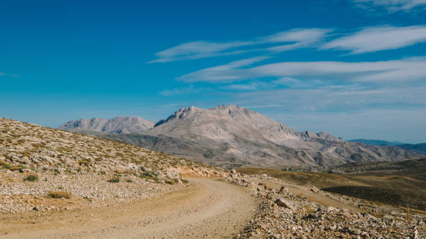 A picturesque autumn landscape features a dirt road winding through the Taurus Mountains, surrounded by vibrant fall foliage. Set at a high elevation, the scene offers breathtaking views of the mountainous terrain, with rolling hills and colorful trees showcasing shades of red, orange, and yellow. The sky above is mostly clear, with a few scattered clouds allowing sunlight to filter through, illuminating the rich colors of the leaves. The dirt road invites exploration, leading viewers deeper into this tranquil outdoor setting, where the beauty of nature and the changing seasons come together in harmony.