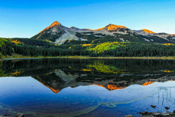 Golden light illuminates rugged mountain peaks reflected in a calm lake, surrounded by lush greenery and forested slopes, captured at sunset in Paonia, Colorado