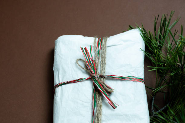 istock Wrapped Christmas gift alongside pine branch on brown surface. 2182112374
