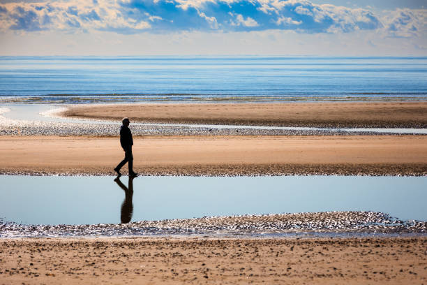 istock Senior man walking on sandy beach alone with calm sea and cloudscape 2182117512