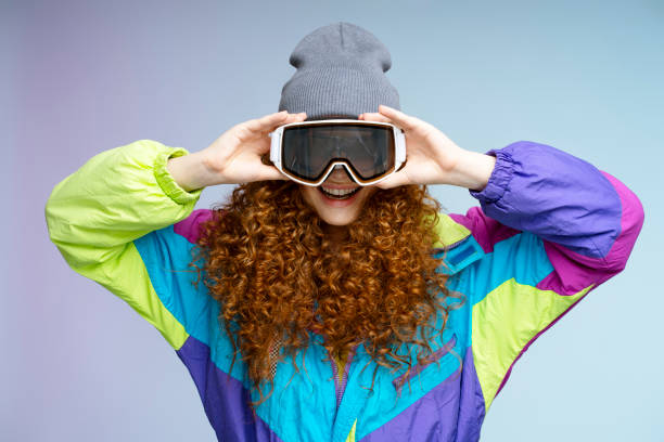 Redhead woman wearing colorful ski suit and gray beanie holding ski goggles and smiling behind them