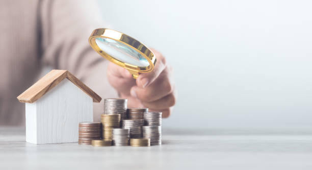 istock Mand holding a magnifying glass over a stack of coins and a house model. The image represents assessing home damage and budgeting for repairs, emphasizing careful financial planning for maintenance 2182119732