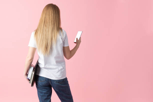 Lifestyle, education, telecommunications, technology concept. A woman holds a phone and a notebook against a soft pink backdrop with copy space. Model standing back to camera