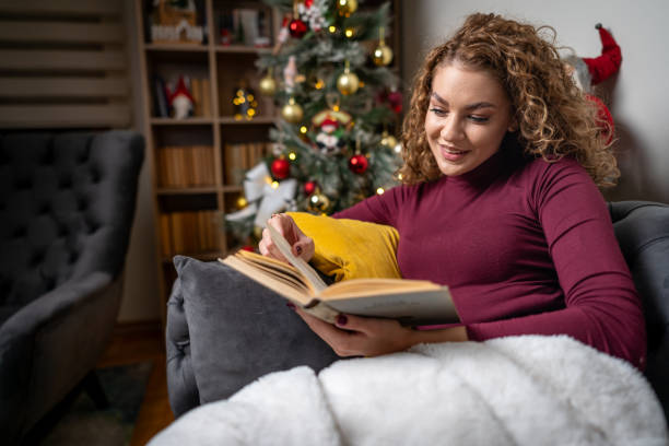istock Young woman lounging on the sofa, under a warm, cozy blanket, reading a book 2182127604