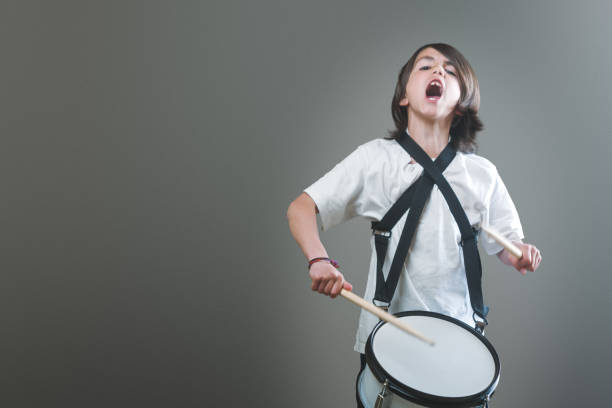 A boy plays a drum with passion isolated on a grey background. He is making a loud noise as he plays