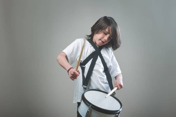 A young boy is playing a drum while wearing a white shirt and black strap. He is focused on his drumming and he is enjoying himself