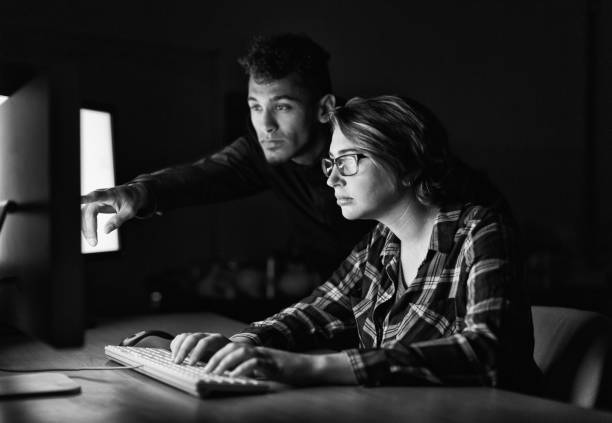 Young man points at computer screen as he collaborates with young woman sitting at desk