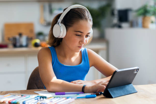 istock Cheerful little girl doing videocall with digital tablet and headphones while doing her homework in the kitchen at home 2182136071