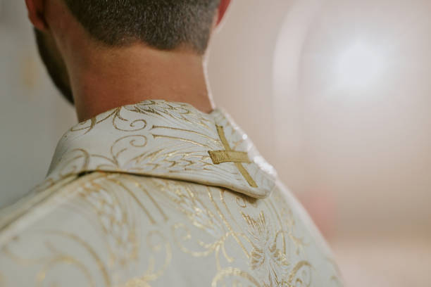 Medium close up of unrecognizable priest wearing white robe with golden embroidered cross and patterns