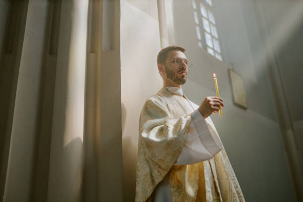 Low angle shot of ample church nave with young Caucasian priest in golden white robe holding long wax candle during service inside