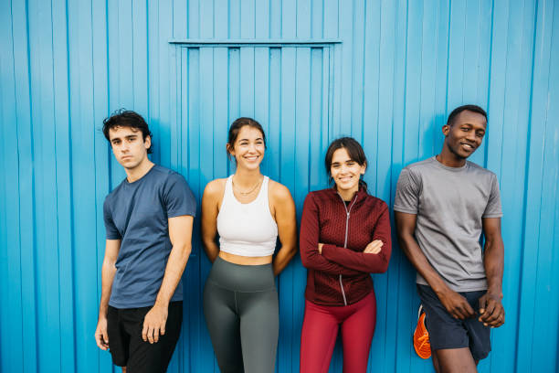 A group of four friends from diverse backgrounds casually leaning against a vibrant blue wall, embodying friendship, diversity, and fitness. They exhibit a relaxed and cheerful demeanor.