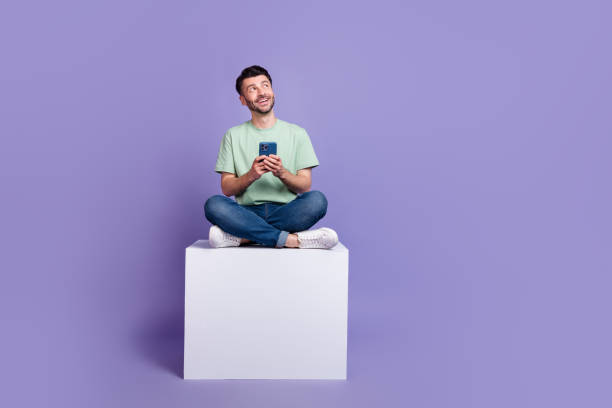 istock Full length photo of nice young man hold gadget look empty space sit cube dressed stylish gray outfit isolated on purple color background 2182152495