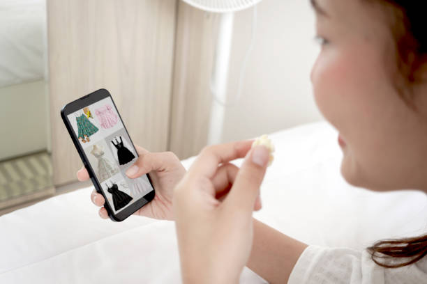 istock Relaxing woman in white bathrobe using mobile phone to shop online while eating popcorn snack and lying on bed at home. Female consumer using smartphone for convenience and versatility purchase process at bedroom. 2182153818