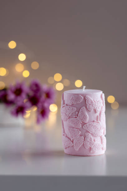 Pink decorative candle with butterflies pattern. Flowers and lights in background with copy space. Home decor. Cozy interior. Soy wax candle making.