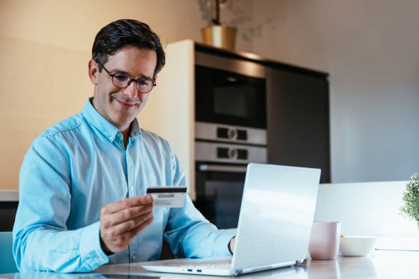 istock Businessman sitting at a kitchen table paying online on a laptop, illuminated by warm light, with a smartphone and credit card 2182157536
