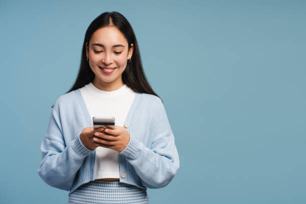 istock Smiling Asian woman holding mobile phone, using mobile app, standing isolated on blue background 2182163295
