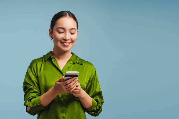 Smiling Asian woman holding mobile phone, using mobile app, standing isolated on blue background