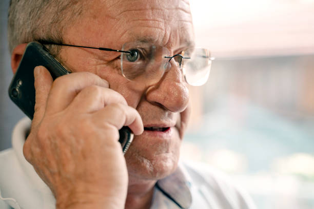 Close up of a senior man wearing glasses making a phone call, holding a mobile phone to his ear, engaged in conversation