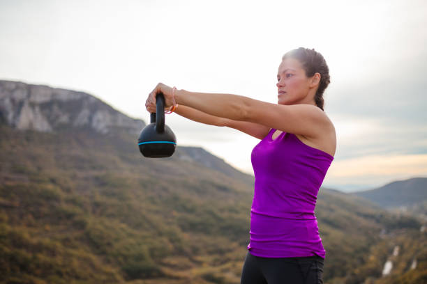 A young woman in athletic wear performs a kettlebell exercise outdoors, surrounded by a beautiful mountain landscape, embodying strength and fitness.