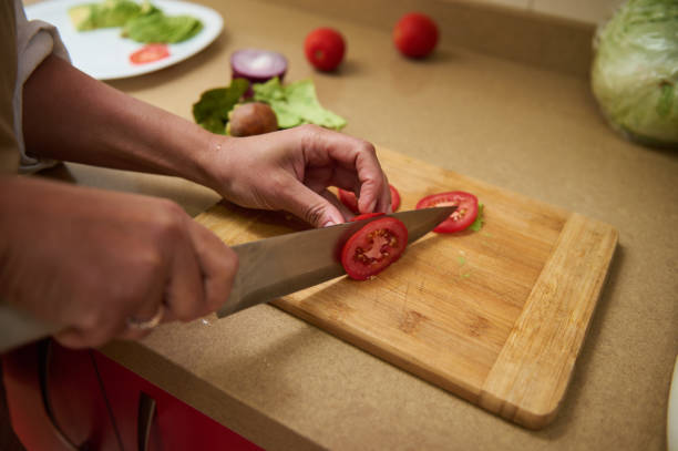A person carefully slices fresh tomatoes on a wooden cutting board, amidst fresh ingredients on the counter, showing culinary preparation.