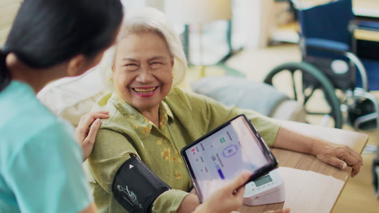 Woman asian nurse taking blood pressure of a senior woman patient at home.Home care healthcare professional using digital tablet